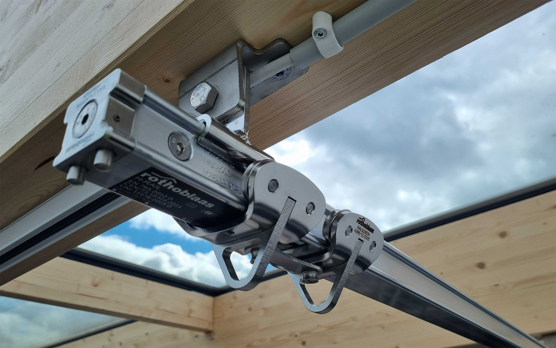 Rothoblaas H-RAIL rigid fall arrest rail system mounted on a glulam timber beam — sliding runner detail on a large-span timber and glass roof structure, Denmark.